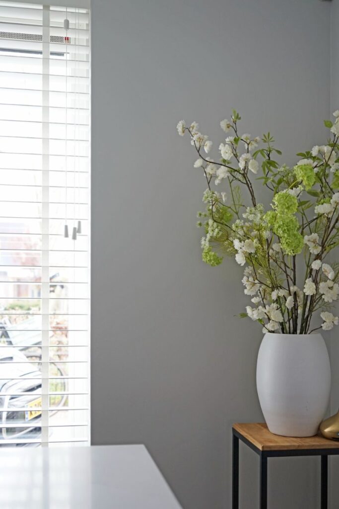 Lichte muur, bijzettafel met bloemen in een vaas. Raam met witte horizontale jaloezieën.