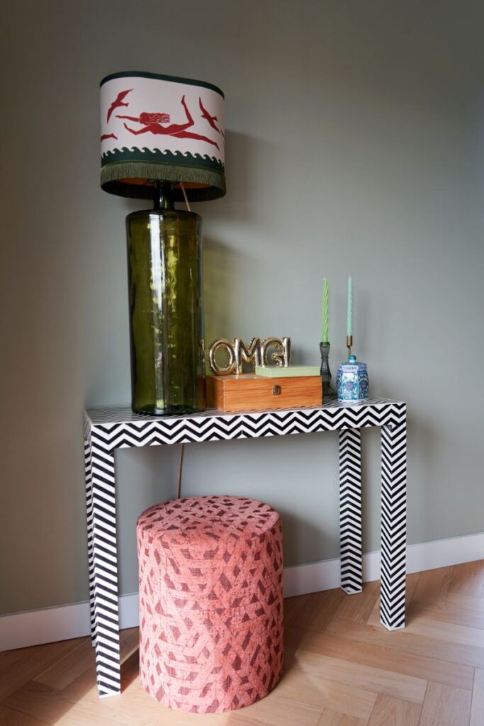 Grijsgroene muur en een visgraat vloer. Sidetable met grote opvallende tafellamp en andere accessoires. Poef in roze stof met een print.