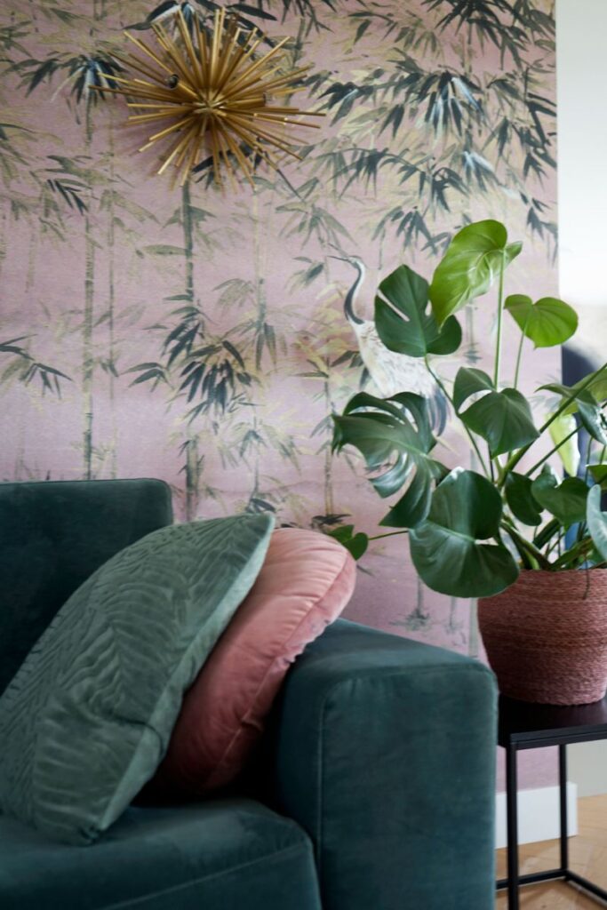 Decoratief print behang met opvallende goudkleurige wandlamp. groene bank met sierkussens en een zwarte bijzettafel met een plant.