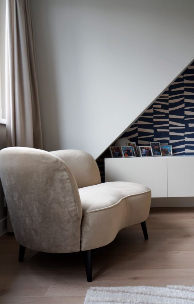 Witte muur en een donkerblauw met wit decoratief behang. Lichte pvc vloer en een wit tv-meubel. Beige chaise longue met zwarte poten.