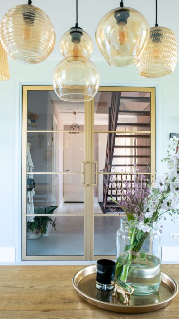 Licht blauwgroene muur met goudkleurige stalen deuren naar de entree. Eettafel met hierboven een hanglamp met verschillende bollen.