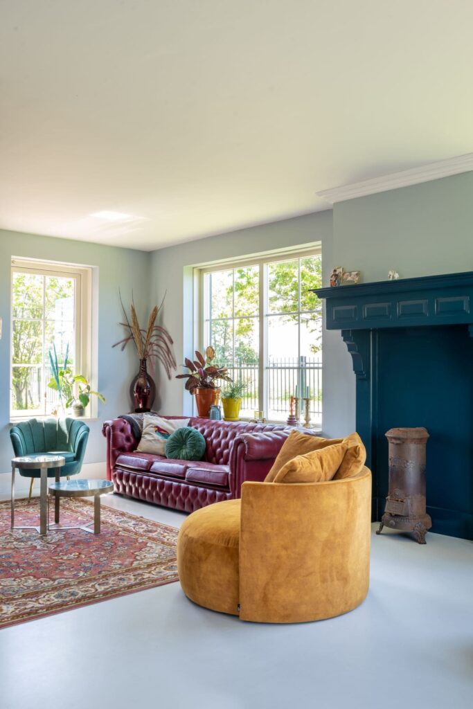 Woonkamer met blauwgroene muren, lichte vloer en plafond. Donkerblauwe schouw. Rode leren chesterfield bank met grote ronde gele fauteuil. Gekleurd vloerkleed met patroon.