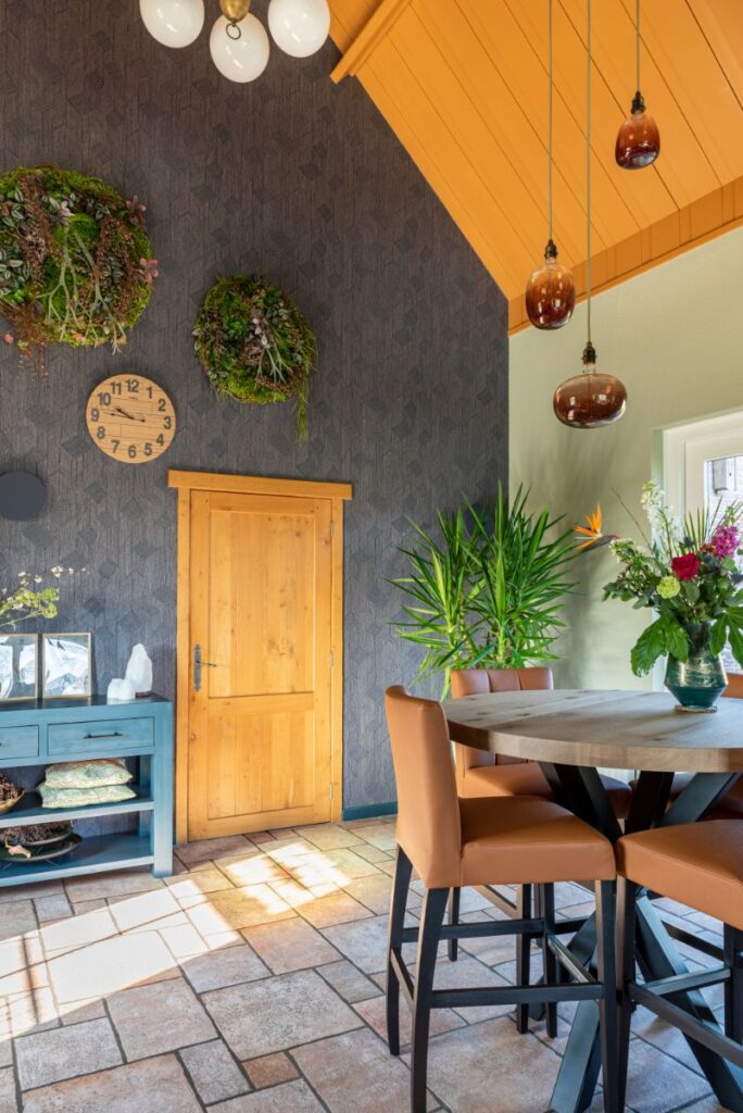 Stenen vloer met groene wand, donkerblauw decoratief behang. Schuine wand in het oker geel. Ronde bar met barstoelen. Planten.