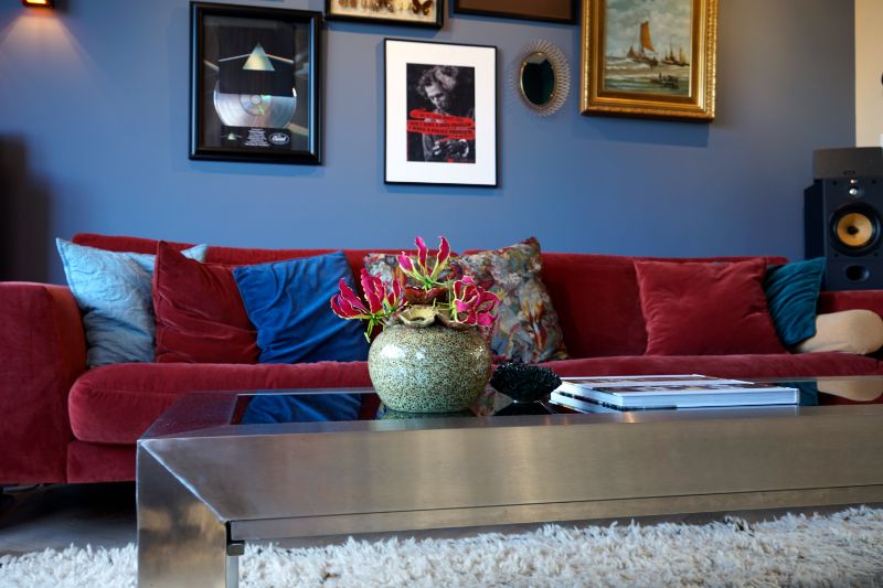 Woonkamer met blauwe muur en een rode bank. Veel verschillende schilderijtjes aan de muur. Metalen salontafel met accessoires.