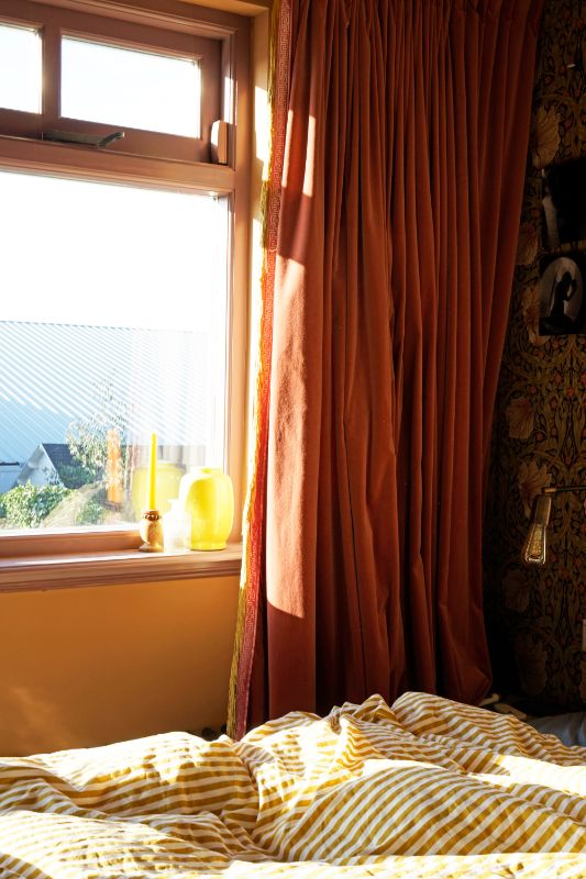 Masterbedroom met decoratief bloemenprint behang. Geel met wit gestreept dekbedovertrek. Rood/oranje gordijnen.