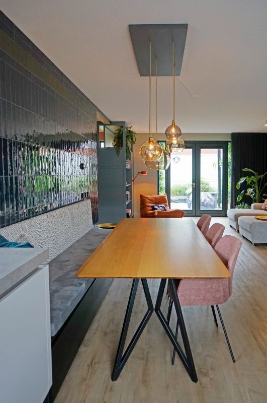 Donkerblauw getegelde muur met een eetkamerbank, tafel, 3 eetkamerstoelen in een roze kleur en mooie glazen lampenbollen.