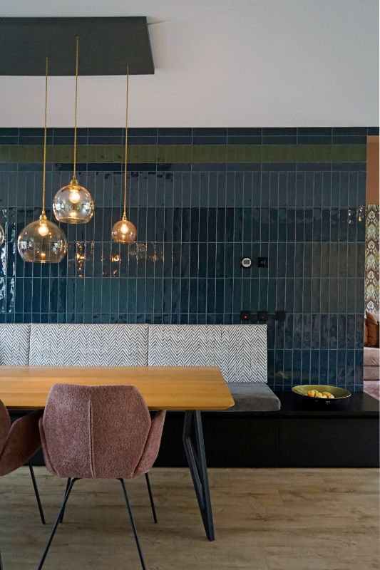 Donkerblauw en groen getegelde muur met een eetkamerbank, tafel, 3 eetkamerstoelen in een roze kleur en mooie glazen lampenbollen.