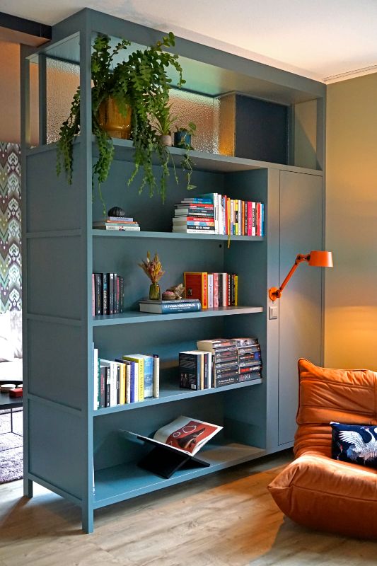 Blauwe kast als roomdivider met boeken en een oranje leeslampje. Hierbij staat een oranje fauteuil.