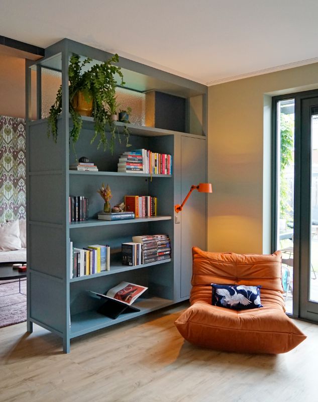 Blauwe kast als roomdivider met boeken en een oranje leeslampje. Hierbij staat een oranje fauteuil.