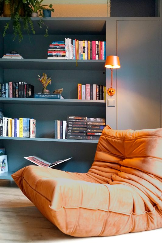 Blauwe kast als roomdivider met boeken en een oranje leeslampje. Hierbij staat een oranje fauteuil.