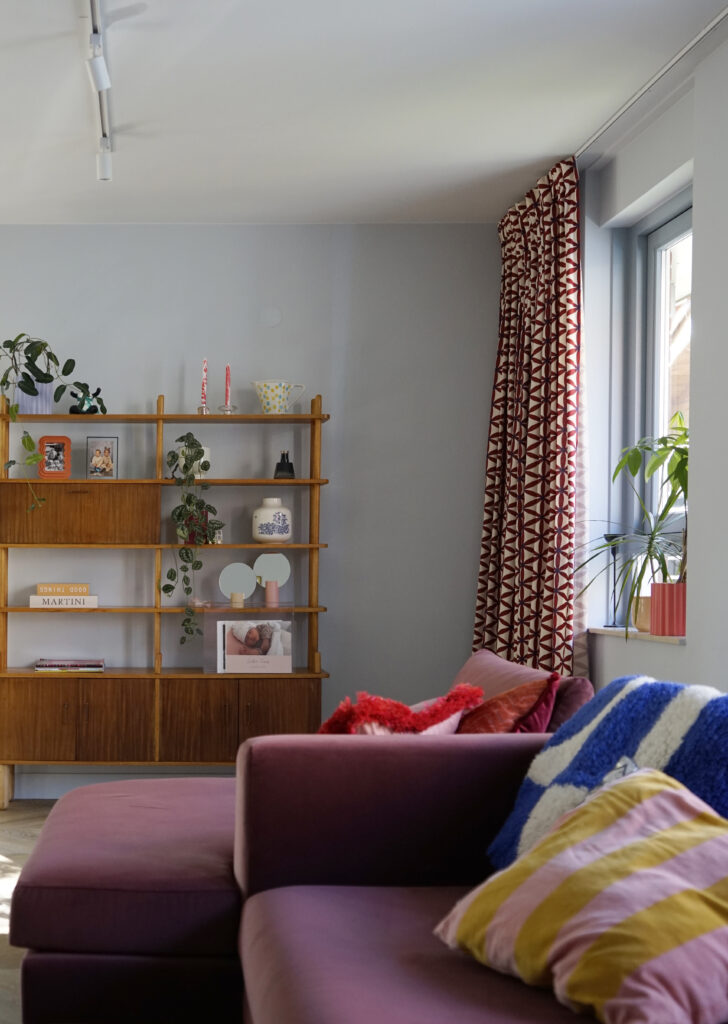 Woonkamer met roze bank en gekleurde kussens. Vintage vakkenkast met gekleurde accessoires.