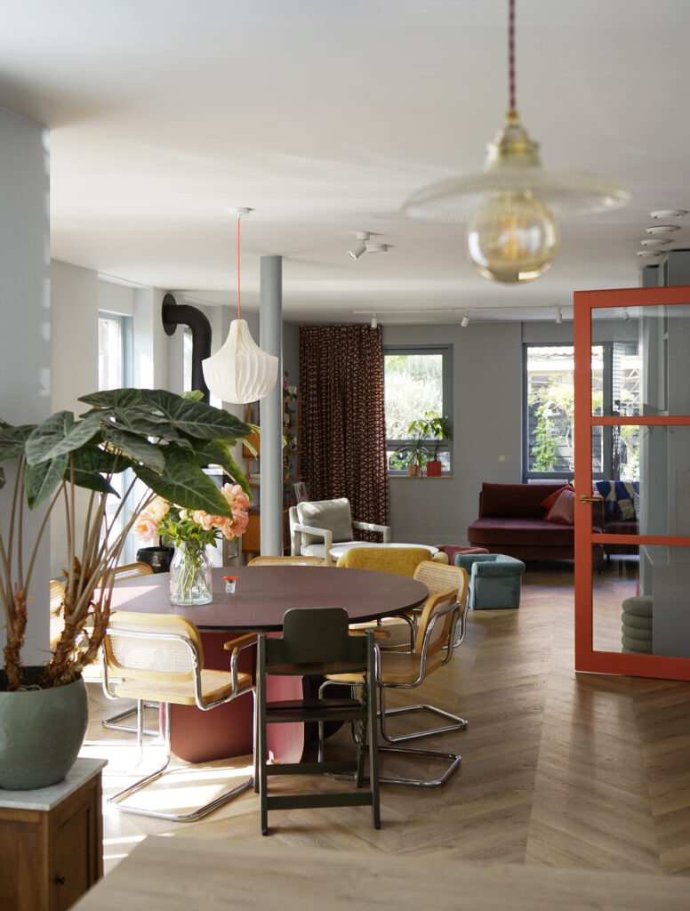 Licht blauwe woonkamer met oranje glazendeur. Ronde bordeaux rode eettafel met rotan stoelen.